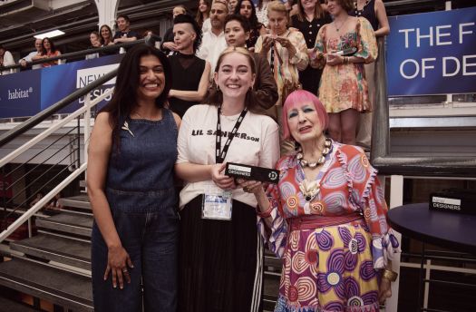 Manchester School of Art student Lil Sanderson in a white t-shirt which says Lil Sanderson in block writing next to fashion designer Zandra Rhodes, a woman with neon pink bobbed hair and a pink dress on. Another woman stands on Lil's left. 